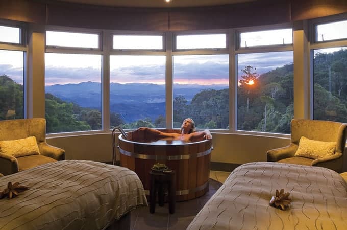 Woman taking a dip on an open bathtub at Lost World Spa in O'Reilly's Retreat in The Gold Coast Hinterland with picture windows and views of the outdoors or surrounding forest during sunset