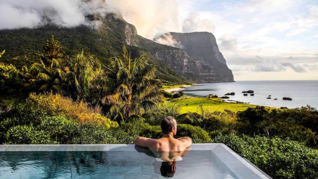 a man on an infinity pool enjoying the views of the surrounding lush greeneries and trees with views of the beach and mountain ranges in the background on the Capella Lodge in Lord Howe Island 