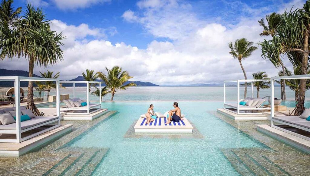 Couple lounging in the middle of a swimming pool on the infinity pool with pool-side beds and seats and palm trees and views of the surrounding beach in InterContinental Hayman Island Resort during daytime