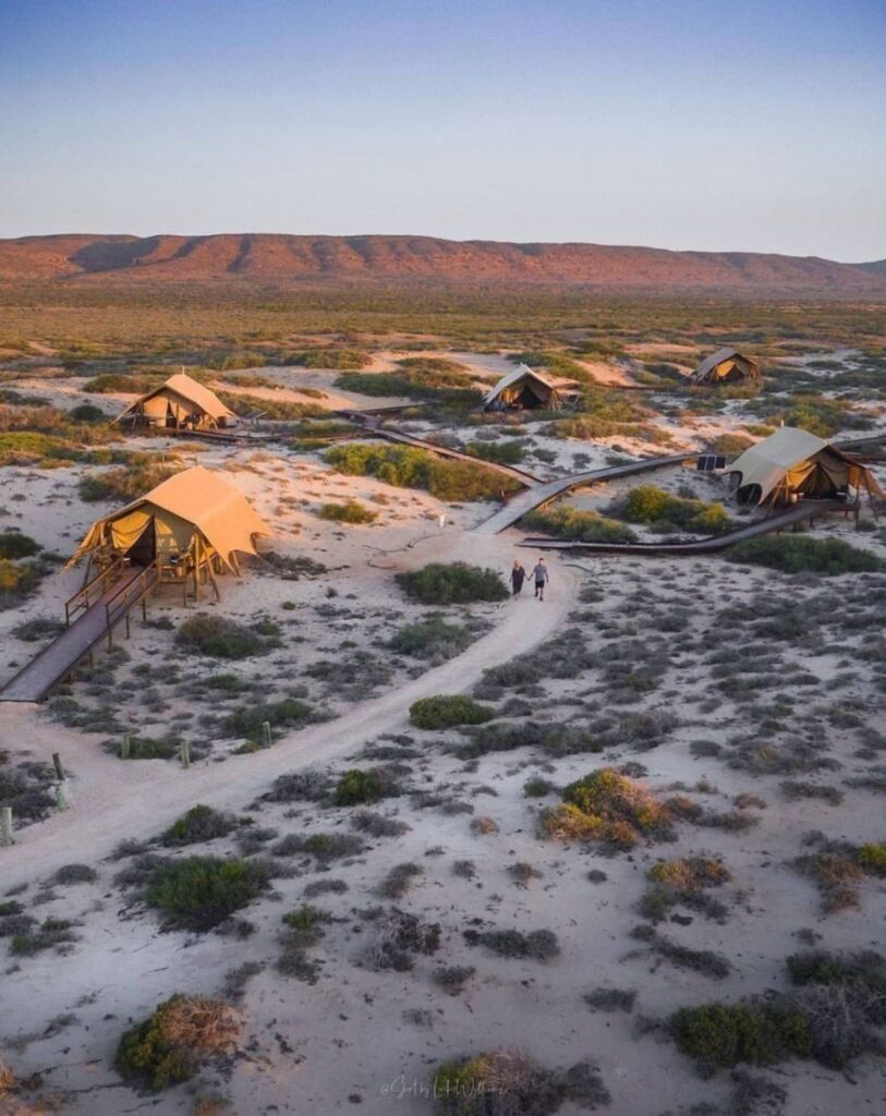 A couple walking on a sandy road with surrounding tents and mountain ranges in the background during sunset on a beach safari camp in Sal Salis Western Australia