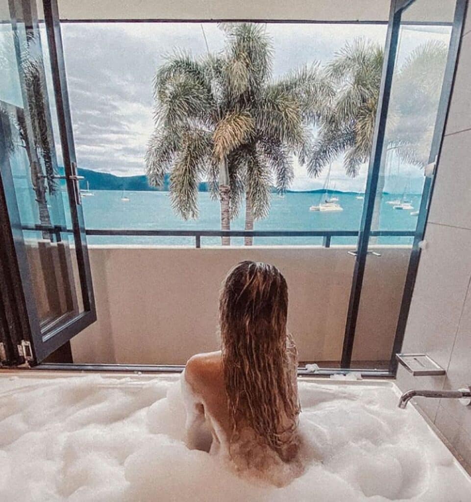A woman taking a bath on a bathtub at the Whitsundays with glass windows and views of the beach