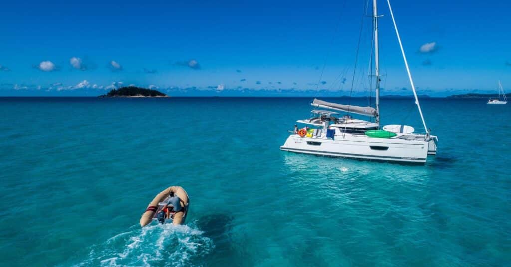 A stationary yacht in the middle of a blue clear water beach with an island in the background and a man riding a raft towards the yacht