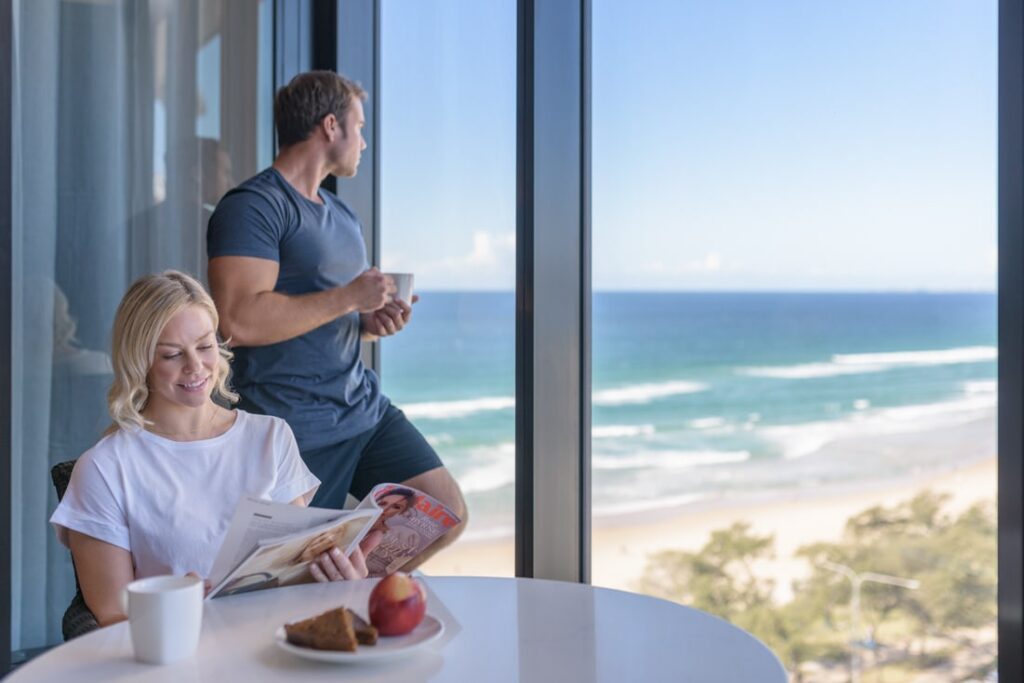 Couple enjoying their breakfast by drinking coffee and reading a magazine at MERITON SUITES SURFERS PARADISE ROMANTIC GOLD COAST ACCOMMODATION with glass walls and views of the beautiful beach