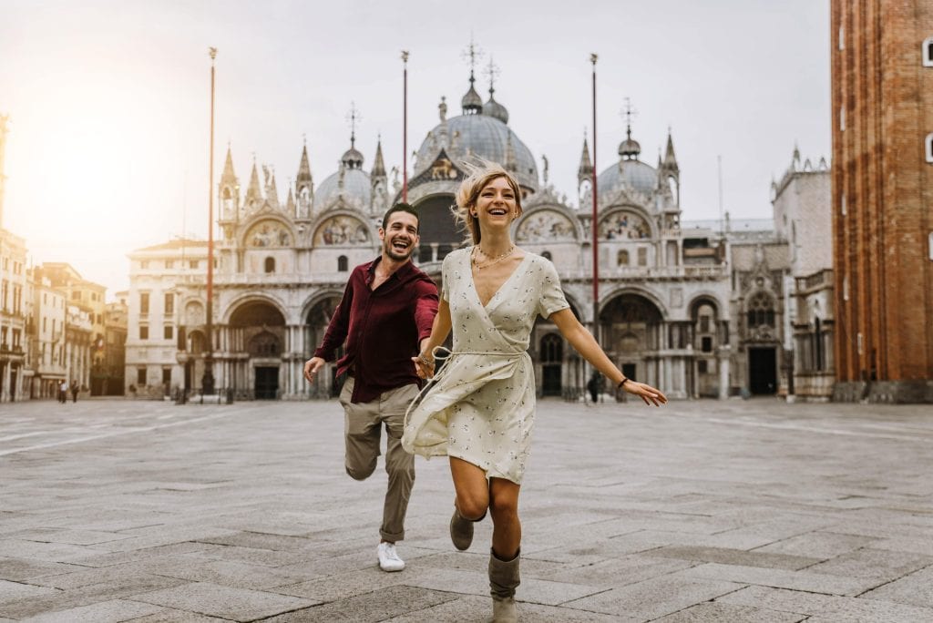 Couple of tourists visiting Venice, Italy - Boyfriend and girlfriend in love running together on city street at sunset - People, love and holidays concept